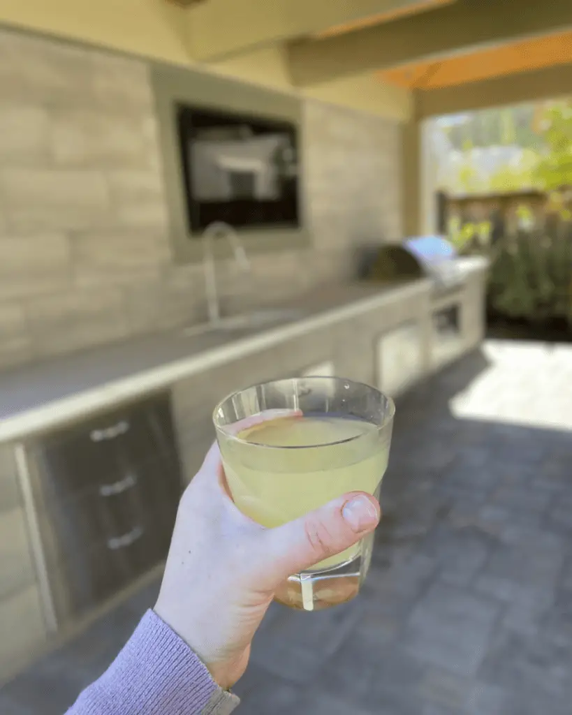 A person holding a glass of yellow liquid, possibly juice, in an outdoor kitchen area with a countertop and grill in the background.