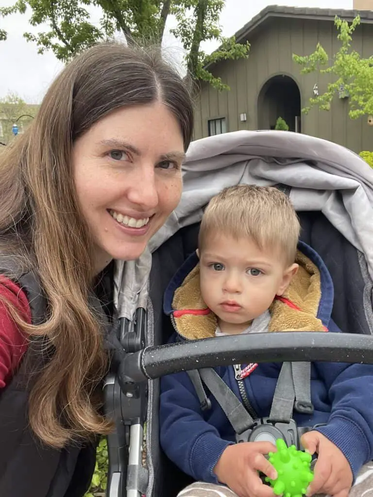 A woman with long brown hair smiles while posing next to a young child sitting in a stroller. The child is wearing a navy blue hoodie with a tan lining and holds a green spiky ball. The background features a building and some trees.