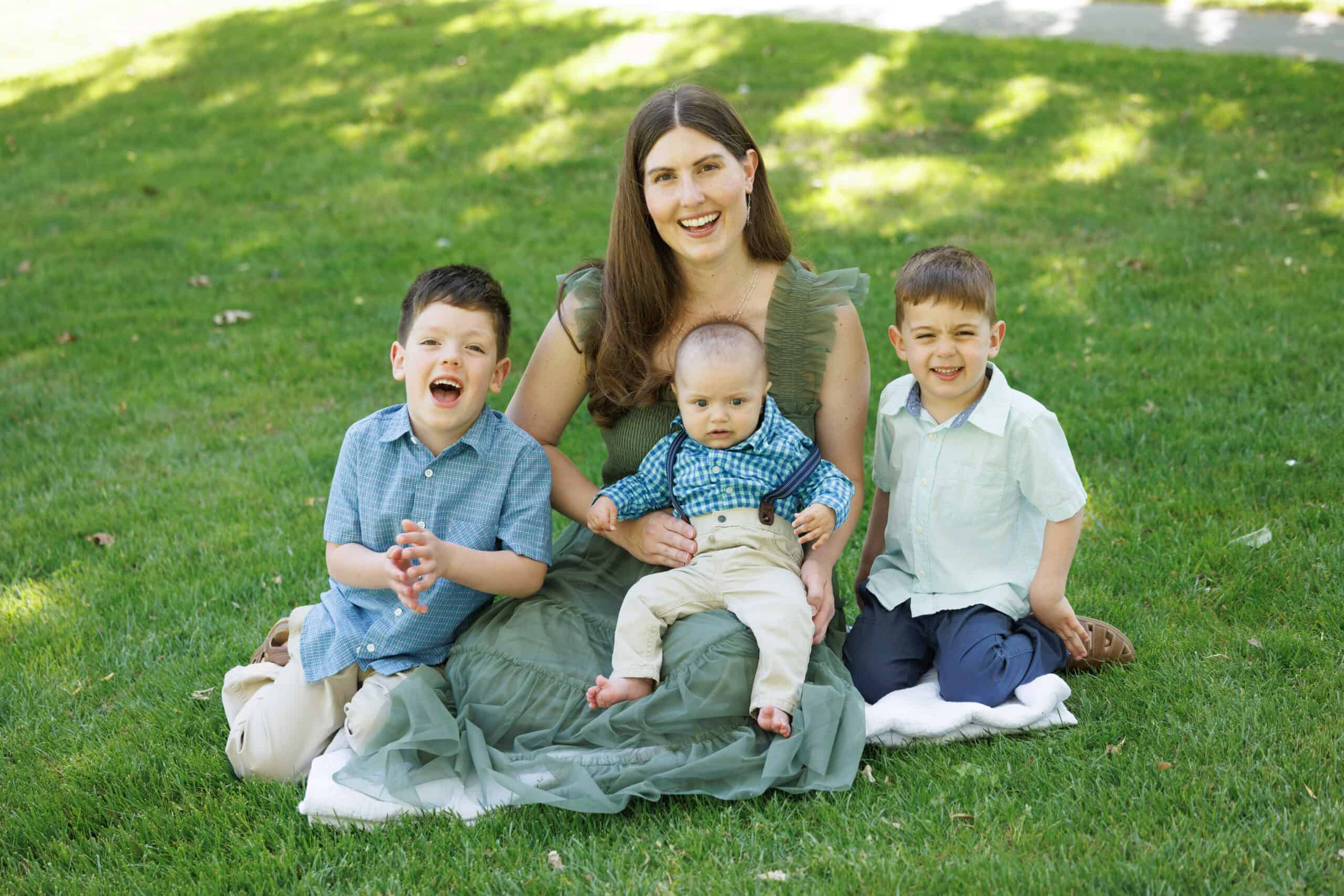 A woman sits on the grass with a baby on her lap and two young boys on either side, all smiling and enjoying a sunny day outdoors.
